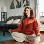Woman with curly hair in an orange sweater sits cross‑legged on a living room rug with headphones, meditating.