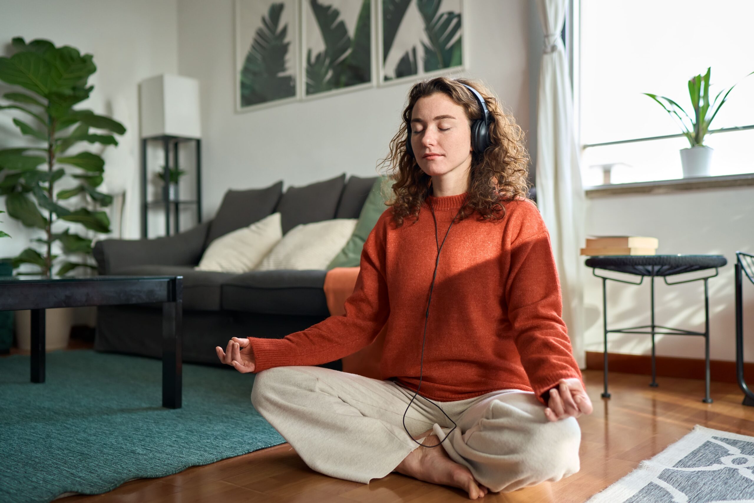 Woman with curly hair in an orange sweater sits cross‑legged on a living room rug with headphones, meditating.