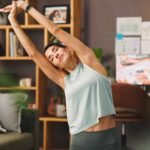 Woman in workout wear stretches arms overhead in a living room, eyes closed, mid-tretch near a sofa and shelves.