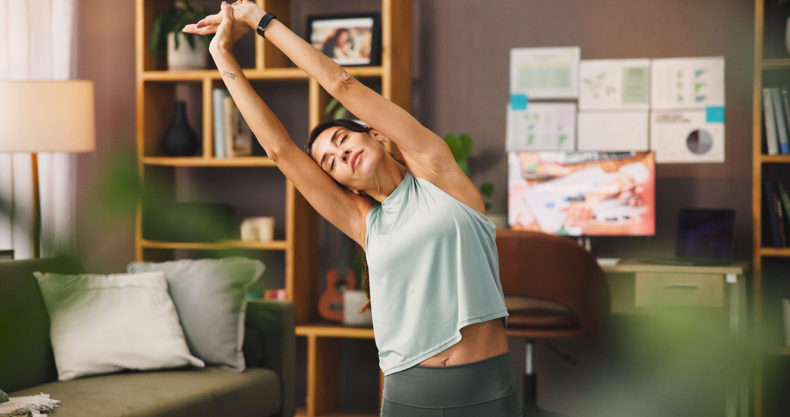 Woman in workout wear stretches arms overhead in a living room, eyes closed, mid-tretch near a sofa and shelves.