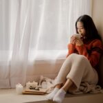 Woman in an orange knit sweater sits by a window, holding a mug, with candles and a notebook on the windowsill nearby.