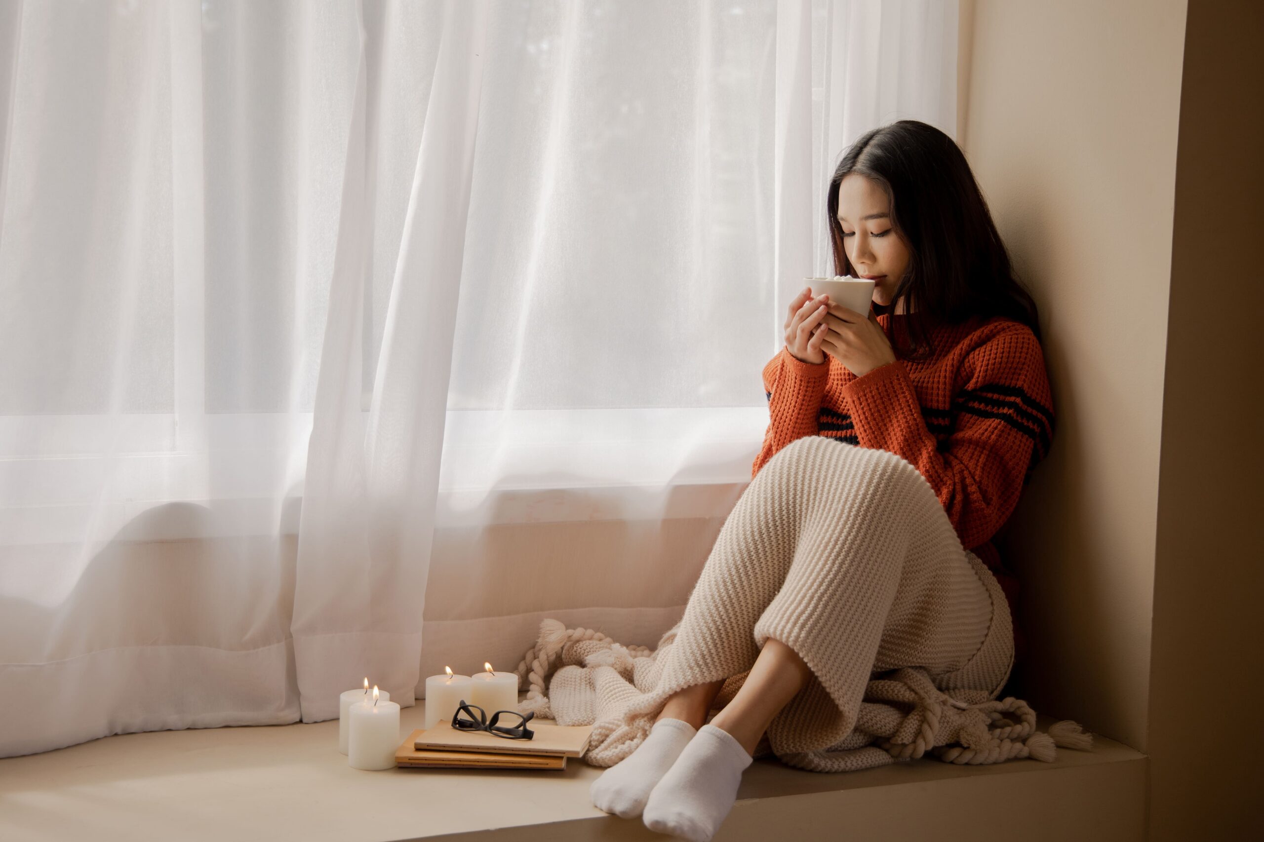 Woman in an orange knit sweater sits by a window, holding a mug, with candles and a notebook on the windowsill nearby.